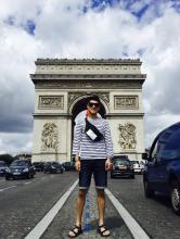 Man in sunglasses, navy/white striped long-sleeved shirt and knee-length jean shorts stands on the center divider of the road leading to the Arc de Triomphe, which perspective makes to appear that he is filling the archway.