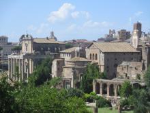 The skyline of rome pictured on a sunny day. Only a few clouds linger above the tan buildings. In the forefront, grass and trees can be seen. 