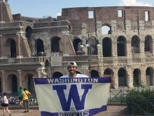 Man with backwards baseball cap holds up UW flag standing in front of the Colosseum in Rome, Italy.
