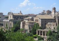 The skyline of rome pictured on a sunny day. Only a few clouds linger above the tan buildings. In the forefront, grass and trees can be seen. 