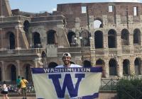 Man with backwards baseball cap holds up UW flag standing in front of the Colosseum in Rome, Italy.