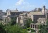 The skyline of rome pictured on a sunny day. Only a few clouds linger above the tan buildings. In the forefront, grass and trees can be seen. 