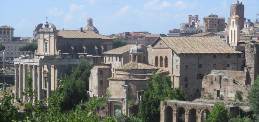 The skyline of rome pictured on a sunny day. Only a few clouds linger above the tan buildings. In the forefront, grass and trees can be seen.